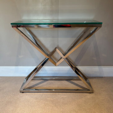 Contemporary Stainless Steel Glass Console Table The Contemporary Stainless Steel Glass Console Table, with a geometric gold metal frame and sleek glass top, stands elegantly against a beige wall on a carpeted floor.