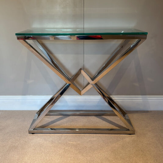 Contemporary Stainless Steel Glass Console Table The Contemporary Stainless Steel Glass Console Table, with a geometric gold metal frame and sleek glass top, stands elegantly against a beige wall on a carpeted floor.