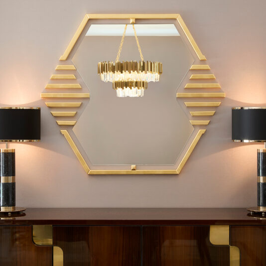 A modern Art Deco-inspired interior features the Art Deco Inspired Hexagonal Mirror with gold accents on a taupe wall. Reflected is a geometric chandelier above a dark wood console with gold details, flanked by two black and gold lamps, adding elegance and charm. Also featuring high-end furniture in the form of a dark wood cabinet