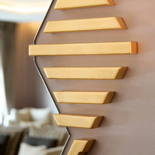 Close-up of an Art Deco design featuring gold bars on a light brown wall with a softly blurred background showing the reflection in an Art Deco Inspired Hexagonal Mirror, glimpsing neutral furniture and curtains.