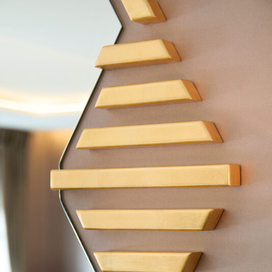 A close-up view of the Art Deco Inspired Hexagonal Mirror showcases a zigzag pattern with gold-colored bars on a smooth, light brown backdrop, blending geometric elements for a modern and stylish aesthetic.
