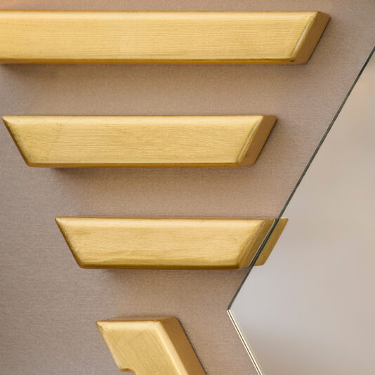 A close-up of a wall showcases geometric patterns with gold textured blocks in diagonal lines on a beige background. The Art Deco Inspired Hexagonal Mirror is visible on the right, introducing a reflective element to the stylish scene.
