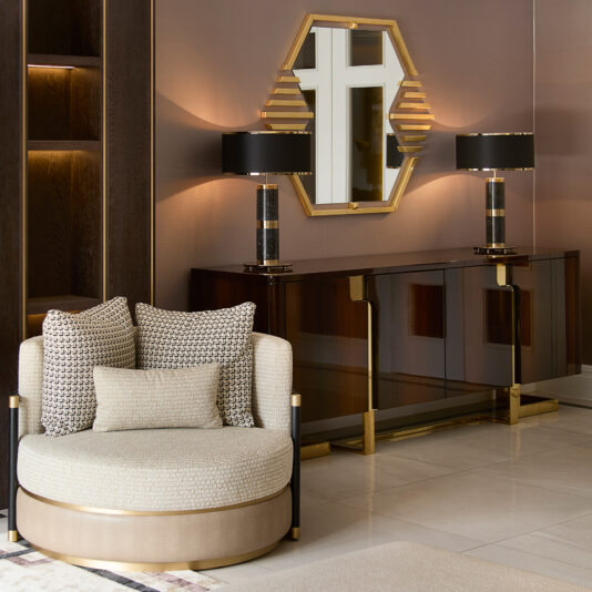 Luxurious living room with a circular beige chair and patterned cushions, a gold-accented wooden sideboard adorned with matching black and gold lamps, topped by the Art Deco Inspired Hexagonal Mirror that enhances ambient light.