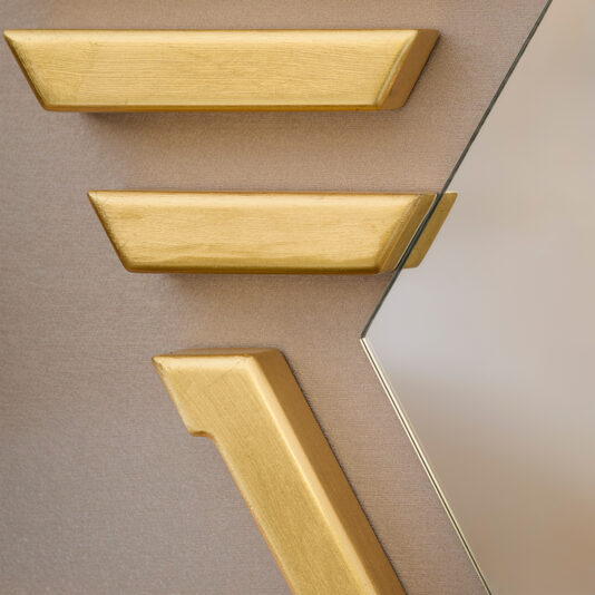 Close-up of the Art Deco Inspired Hexagonal Mirror partially reflecting a wall, featuring three gold geometric wooden pieces on a light brown textured surface, creating an abstract visual against the muted background.