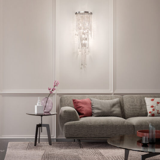 A modern living room showcases a gray couch with assorted pillows, a side table with a pink flower vase, and an Oval Cut Crystal Pendant Chandelier elegantly illuminating the space. A patterned rug adds texture to the light-colored floor.