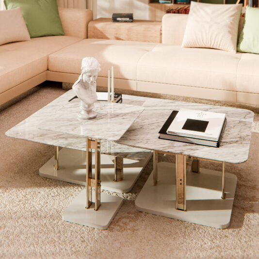 A Round Modern Marble Coffee Table elegantly rests on a plush rug in the living room, displaying a small bust sculpture, books, and candles. Cream-colored sectional sofas with green and beige cushions encircle the table, creating a cozy atmosphere.