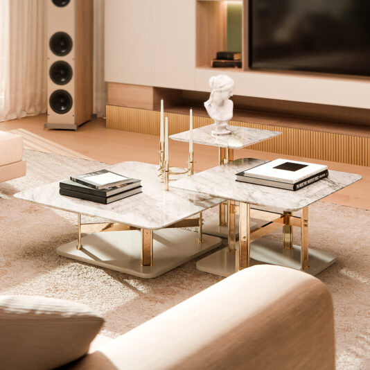 A modern living room showcases the Contemporary Marble Coffee Table Set, adorned with books, a small statue, and golden candle holders. The area includes a plush rug, beige couches, and a sleek speaker near a large TV, creating an elegant and luxurious ambiance.