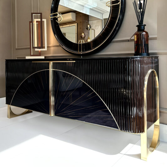 The Luxury Art Deco Style Sunburst Sideboard in black and gold with vertical lines and curved gold accents stands against a beige wall, topped with a brown vase, black reed diffuser, and a large round mirror reflecting the ceiling.