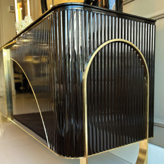 The Luxury Art Deco Style Sunburst Sideboard features a glossy black ribbed surface, gold metal accents, and a large arch detail on the side, shown here on a light floor against a neutral wall.