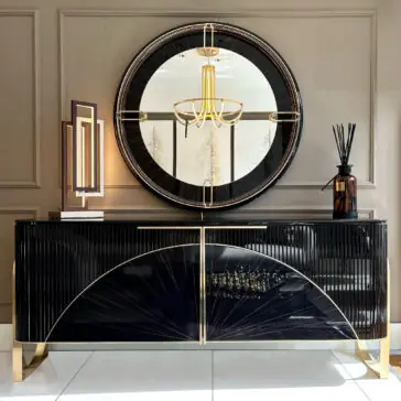 Luxury Art Deco Style Sunburst Sideboard The Luxury Art Deco Style Sunburst Sideboard, finished in glossy black with gold accents, stands against a taupe wall. A large round mirror above reflects a gold chandelier, while decor like a lamp and diffuser adorns the sideboard.
