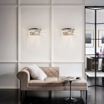 Modern Smoked Cut Crystal Wall Light Elegant room featuring a beige chaise lounge with a white pillow, a small round black table with a magazine, and two decorative wall lights casting patterns on a paneled wall. A Modern Smoked Cut Crystal Tiered Chandelier adds glamour, while glass doors reveal artwork and vases.