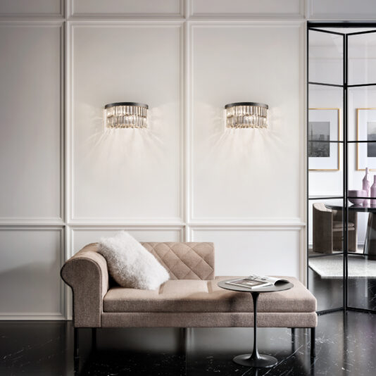 Modern Smoked Cut Crystal Wall Light Elegant room featuring a beige chaise lounge with a white pillow, a small round black table with a magazine, and two decorative wall lights casting patterns on a paneled wall. A Modern Smoked Cut Crystal Tiered Chandelier adds glamour, while glass doors reveal artwork and vases.