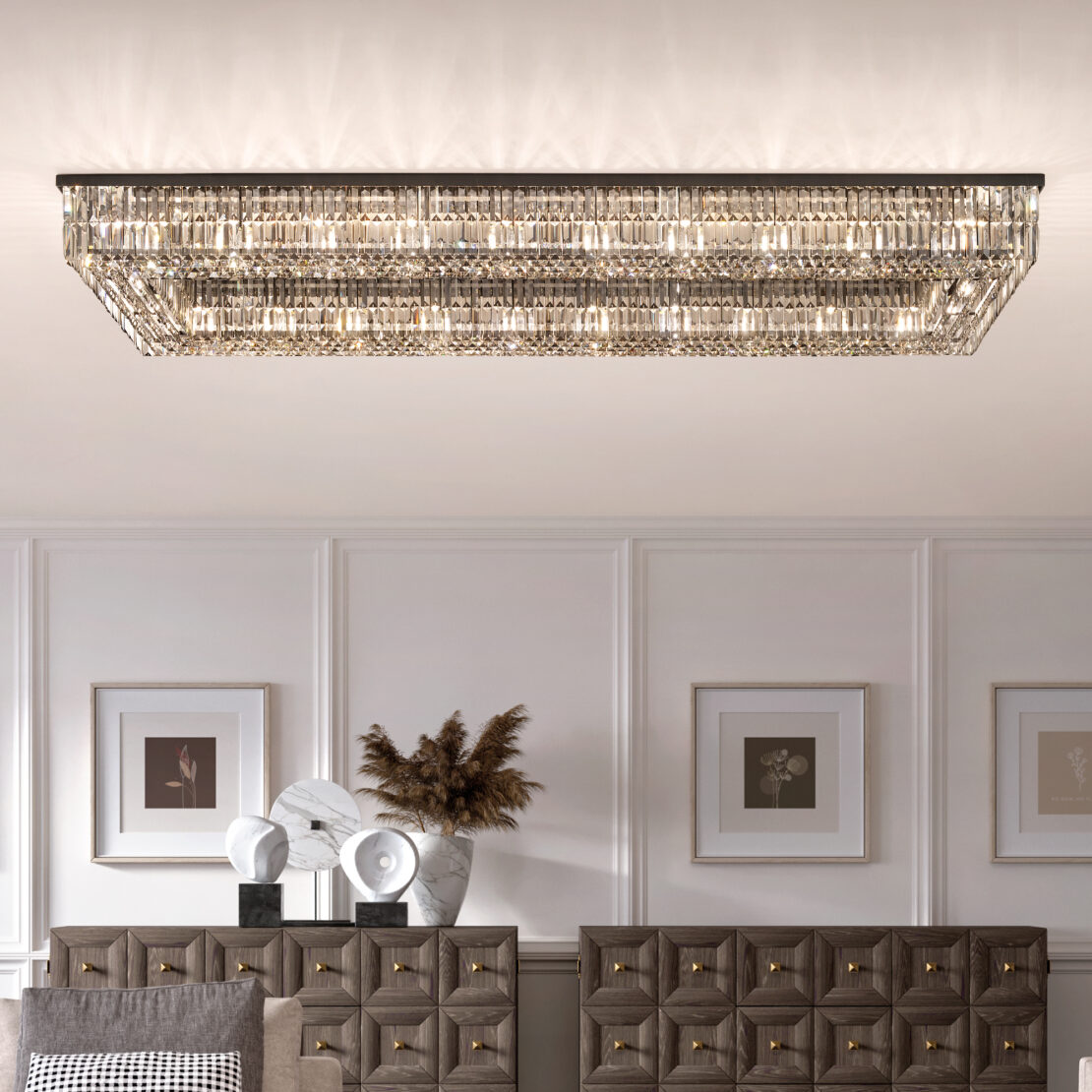 A Modern Oval Smoked Cut Crystal Chandelier graces the white ceiling, casting a sophisticated glow over a room adorned with framed art, while gray tufted furniture and decorative sculptures with gold accents add an elegant touch.