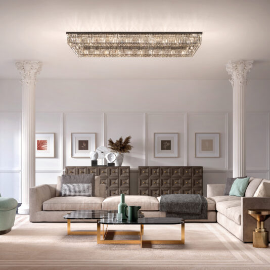 A chic living room features a large beige sectional sofa, glass coffee table, and modern artwork. The ceiling showcases a Modern Oval Smoked Cut Crystal Chandelier. Plush pillows and a blanket add comfort, with elegant columns seamlessly flanking the space.