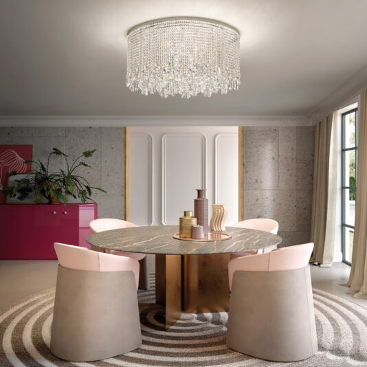 In a contemporary dining room, a round marble table is paired with four beige and pink chairs. The Rectangular Smoked Cut Crystal Ceiling Light elegantly hangs above. A pink sideboard with plants and geometric art decorates the back wall, while light curtains drape over the large windows.