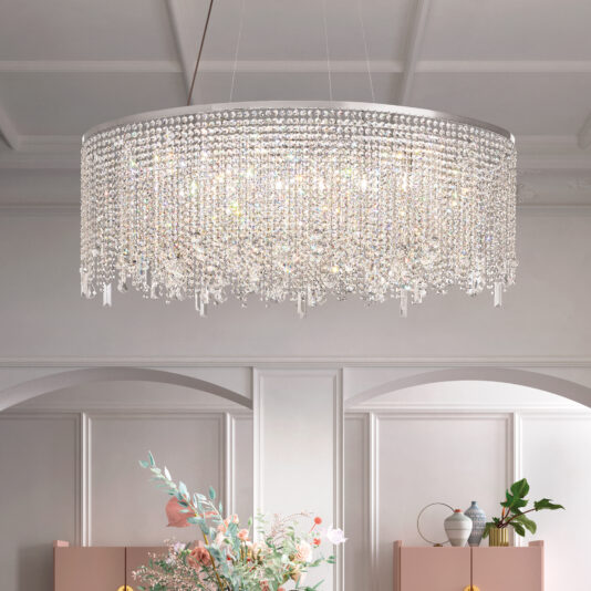 A luxurious Oval Cut Crystal Pendant Chandelier hangs from the ceiling, emitting a bright glow with cascading crystal rows. Below, a room with white-paneled walls showcases a flower arrangement and decorative vases on a shelf.
