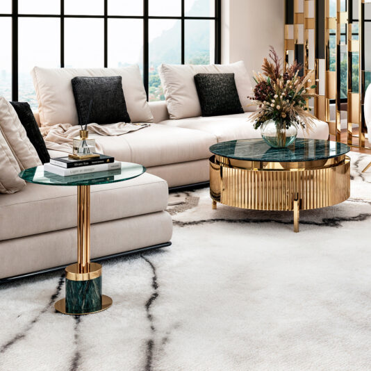 A luxurious living room includes a white sofa with black pillows, a glass round side table with books and a lamp, and the modern Round Modern Marble Coffee Table showcasing flowers—all on a stylish white and black patterned rug under large windows.