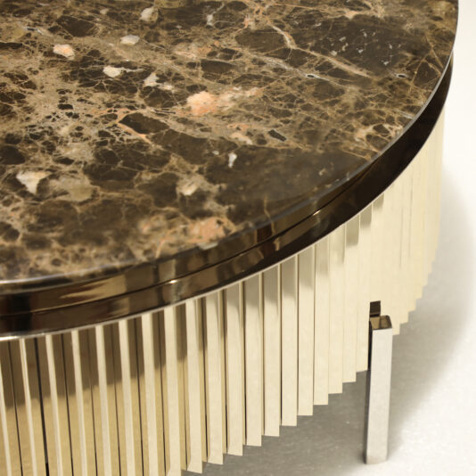 Round Modern Marble Coffee Table The Round Modern Marble Coffee Table features a polished brown marble top with natural patterns, set on a gold metallic base with vertical ridges. It stands out against a plain background, showcasing its sleek and contemporary design.