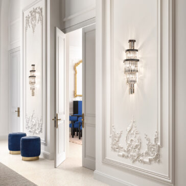 Smoked Cut Crystal Tiered Wall Light An elegant interior with ornate white walls features intricate moldings and a Smoked Cut Crystal Tiered Wall Light. An open double door reveals blue chairs and a large gold-framed mirror. Two round blue ottomans rest in the foreground on a speckled floor.