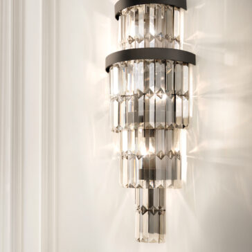 Smoked Cut Crystal Tiered Wall Light The Smoked Cut Crystal Tiered Wall Light features multiple faceted glass tiers and a metal frame. When illuminated, it casts reflections and patterns on the white wall, enhancing its modern design.