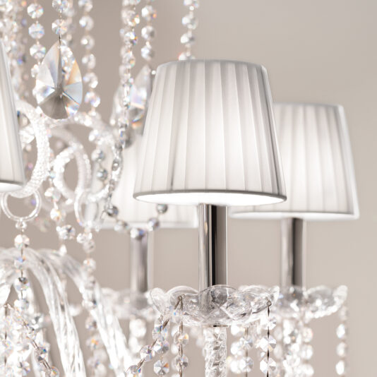 Close-up of the Classic Style Clear Crystal Chandelier with beads, prisms, metallic candle-style holders, and elegant pleated white lampshades casting a soft glow. The softly blurred background enhances its timeless allure.