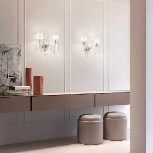 A modern, minimalist vanity with a long taupe countertop, two round taupe stools, decorative vases, abstract wall art, and Classic Style Clear Crystal Wall Lights with pleated white shades.