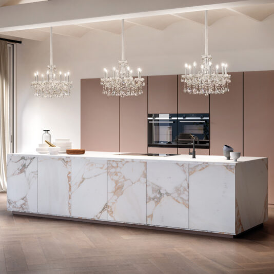 Classic Style Crystal Chandelier A modern kitchen with a large marble island, muted-tone minimalist cabinets, built-in stainless steel appliances, wood flooring, and two Classic Style Crystal Chandeliers overhead.