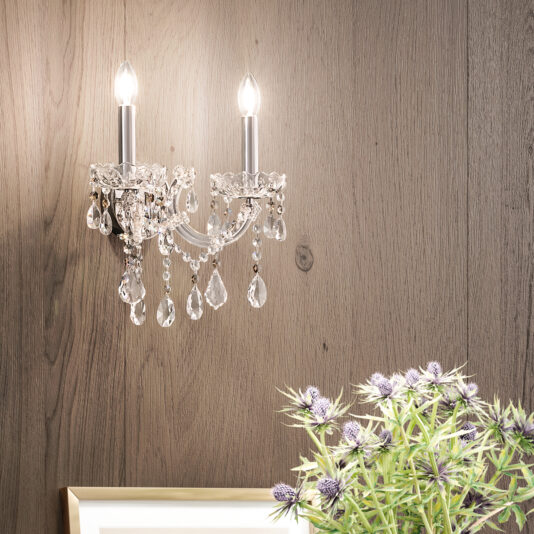 A crystal wall sconce with two candle-shaped bulbs is mounted on a wooden wall, reflecting the elegance of the Oval Crystal Ceiling Light (Copy). Below, a framed picture and purple thistle flowers are partially visible.