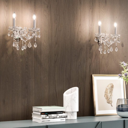 Two crystal wall sconces with exposed bulbs are mounted on a wood-paneled wall above a minimalist console table, complementing books, a white sculpture, and a framed gold leaf print under the Oval Crystal Ceiling Light (Copy).