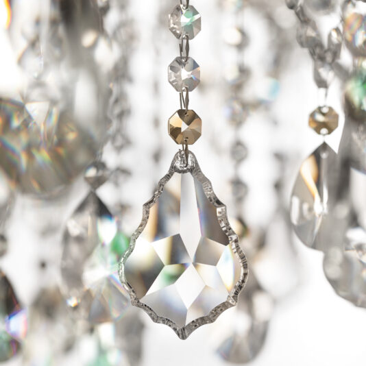 A close-up of the Oval Crystal Ceiling Light (Copy) highlights a teardrop crystal reflecting rainbow light, while other blurred crystals in the background create a sparkling, elegant atmosphere.