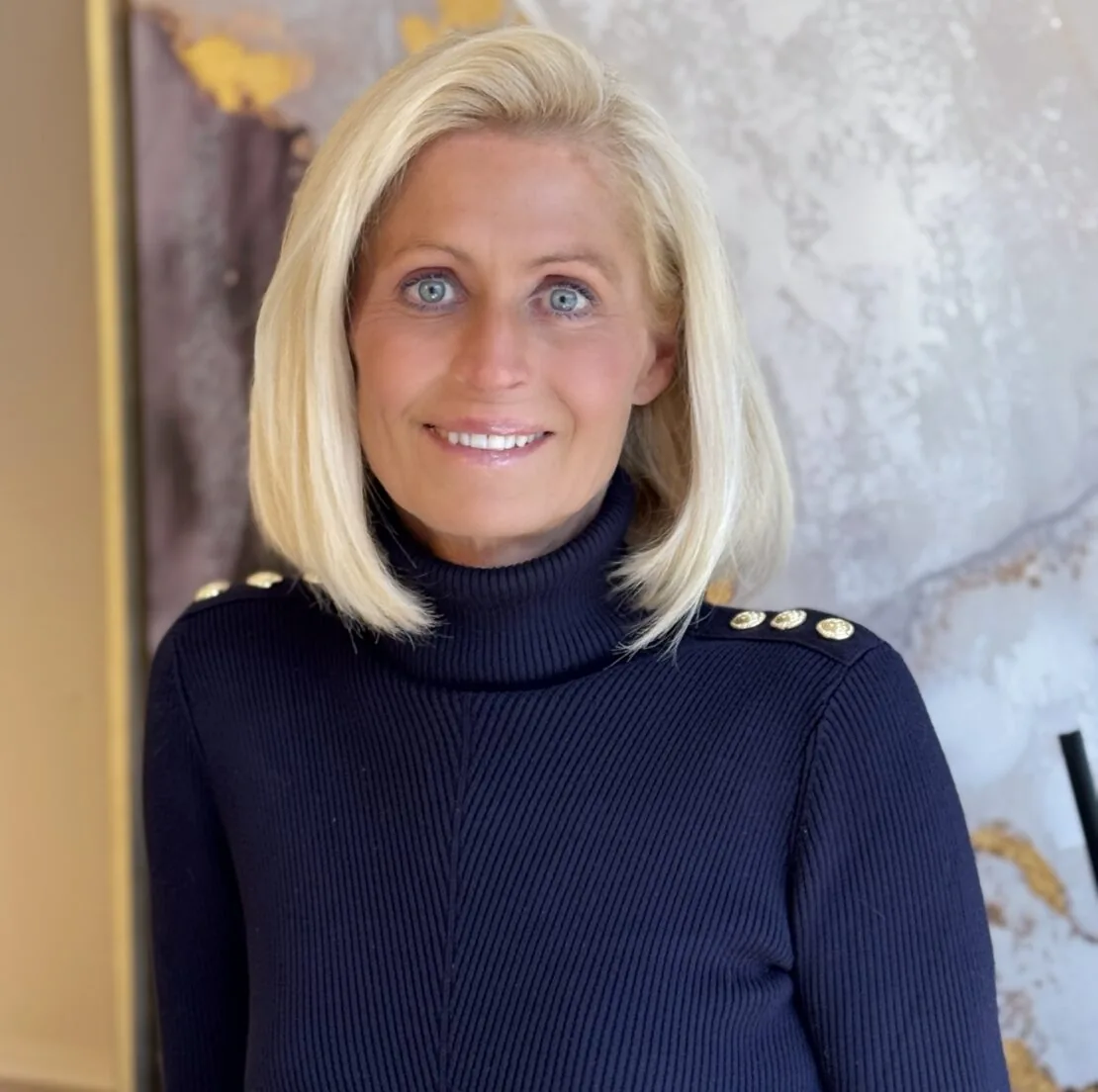 A woman with straight blonde hair and blue eyes smiles at the camera. A luxury interior designer, she wears a navy blue ribbed turtleneck with gold buttons. The background is a softly blurred abstract artwork.
