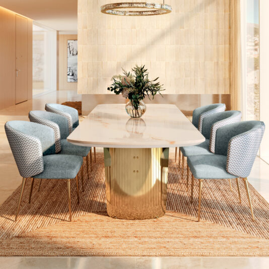 The Large Modern Marble Dining Set centers the room, featuring an elegant table with a gold cylindrical base and eight blue upholstered chairs. A charming vase of flowers sits atop, while beige walls, a sunlit window, and an overhead chandelier add warmth and a welcoming glow.