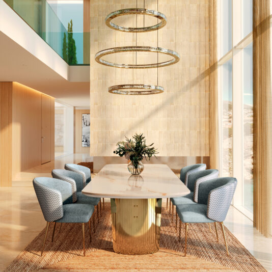 A modern dining space features a large marble table from the Large Modern Veneered Dining Set, surrounded by eight blue upholstered chairs. Three circular chandeliers provide illumination, a floral vase centers the table, and tall windows with a neutral palette enhance the elegance.