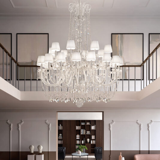 Large Tiered Crystal Chandelier The Tiered Classic Style Clear Crystal Chandelier (Copy) with multiple white lampshades hangs in an elegant double-height room featuring classic molding, a balcony, framed artwork, and bookshelves.