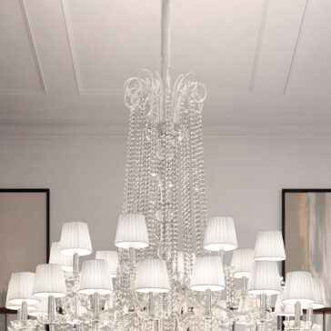 A close-up of the Tiered Classic Style Clear Crystal Chandelier (Copy) with white lamp shades, sparkling beads, and ornate details hangs from a high white ceiling, with parts of two framed artworks visible behind it.