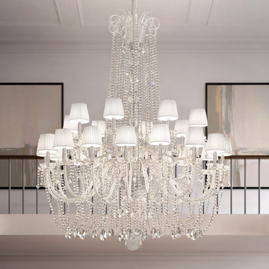 The Tiered Classic Style Clear Crystal Chandelier (Copy) features multiple arms and white lampshades, hanging from a high ceiling in a modern room with abstract art and a balcony railing in the background.