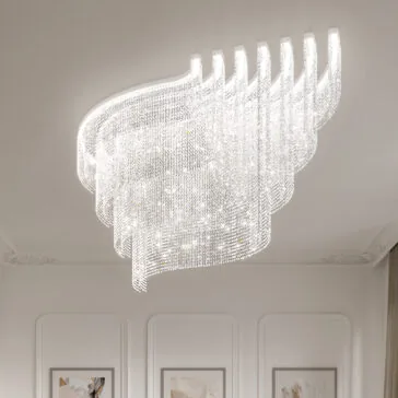 Large Tiered Cut Crystal Ceiling Light The Large Oval Cut Crystal Ceiling Light, featuring an elegant modern wavy design, hangs from a white ceiling. Its chandelier glimmers, with the room’s white walls showcasing molding and framed art.