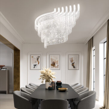 Large Tiered Cut Crystal Ceiling Light A modern dining room showcases a large rectangular black table with gray chairs around it. The Large Oval Cut Crystal Ceiling Light hangs above. Abstract art decorates the walls, tall windows are dressed with beige curtains, and a vase of pampas grass adorns the table.