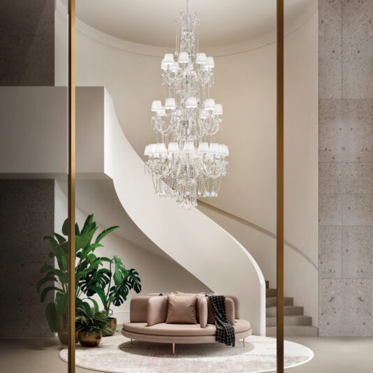 A modern lobby showcases a round beige sofa with cushions and a dark throw, a large potted plant, a circular rug, and the Large Tiered Crystal Chandelier (Copy) beneath a sweeping curved staircase.
