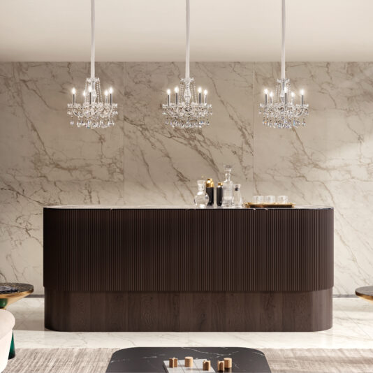 A modern bar with a dark wood finish stands before a marble wall, topped with glassware. Three elegant crystal chandeliers and the Ornate Crystal Wall Light cast a soft glow over the space.