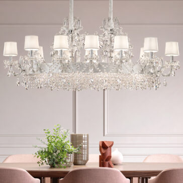 A Round Silver Leaf Crystal Chandelier (Copy) with multiple lampshades hangs above a modern dining table set with pink chairs, a green plant, a vase, and a wooden sculpture. Elegant white wall paneling enhances the sophisticated scene.