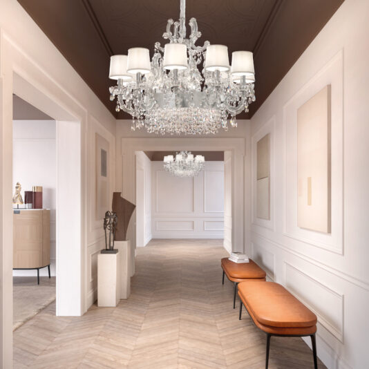 A bright, elegant hallway with white walls, herringbone wood floors, two orange benches, modern art, and two Classic Style Clear Crystal Chandeliers (Copy) hanging from the ceiling.