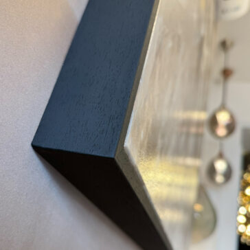 Contemporary Abstract Original Artwork Close-up of the corner of a framed Contemporary Abstract Original Artwork with a black wooden edge, photographed at an angle. Blurred hanging lights and interior elements appear in the background.