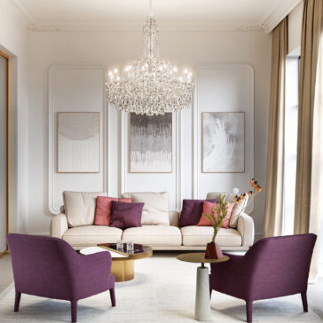 Elegant living room with a white sofa, purple armchairs, and abstract art. A Traditional Style Crystal Chandelier (Copy) hangs from the ceiling while large windows with beige curtains brighten the space.
