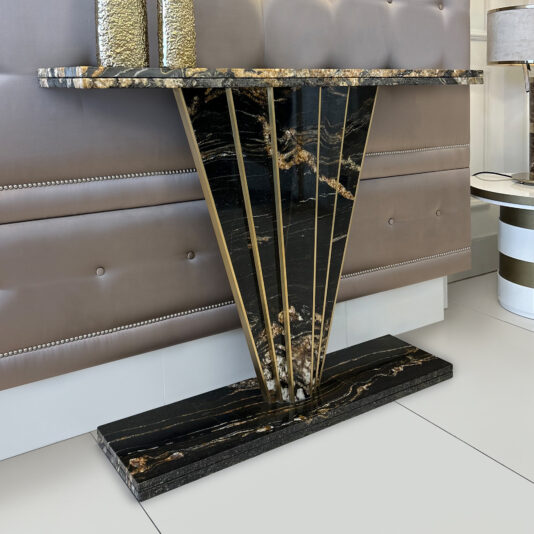The Exclusive Art Deco Style Console Table features a black marble top and base, gold accents, and an angular V-shaped support, set against a beige upholstered wall with decorative studs.