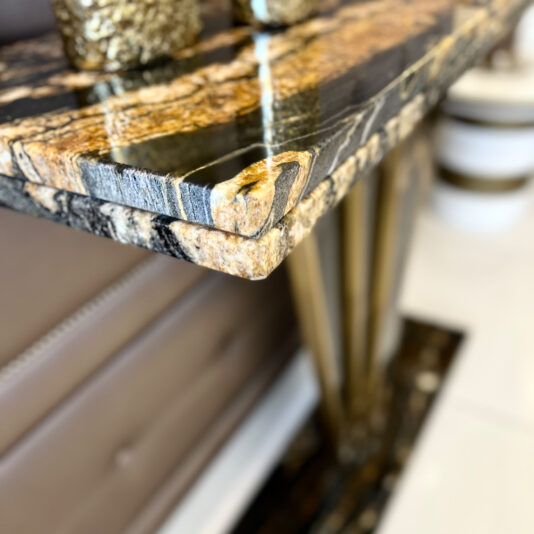 Close-up of the Exclusive Art Deco Style Console Table’s polished marble corner with black, gold, and beige veining reflecting light. A blurred sofa and floor in the background enhance its luxurious appeal.