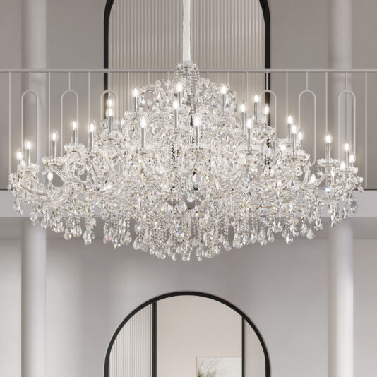 A Large Clear Crystal Chandelier (Copy) with multiple lit bulbs hangs in a bright, elegant room featuring white walls, stately columns, and an arched doorway.