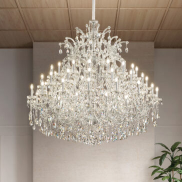 The Large Clear Crystal Chandelier, sparkling with dangling crystals and many lit bulbs, hangs from the wooden ceiling of an elegant room, illuminating the space while a green plant adds grace to the corner.