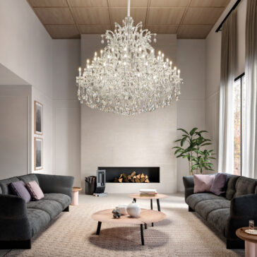 A modern living room with two dark gray sofas, a round wooden coffee table, a Large Clear Crystal Chandelier, a fireplace with stacked firewood, framed art on the wall, and a potted plant near tall curtained windows.