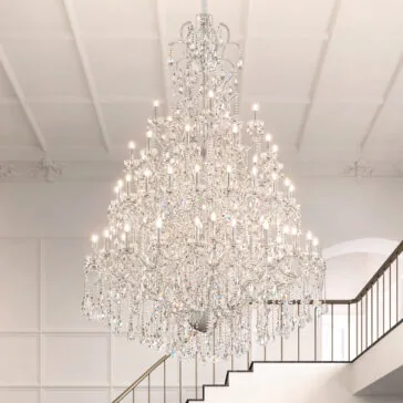 Large Statement Crystal Chandelier The Grand Crystal Crystal Chandelier (Copy) hangs from a high ceiling in a majestic interior with white walls and a staircase to the right, illuminating the space with its many brilliant lights.